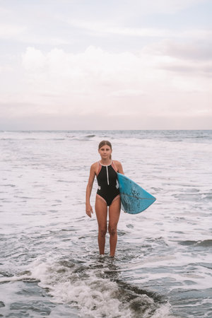 A teenage girl stands in a swimsuit in the ocean and holds surfboard in her handの写真素材