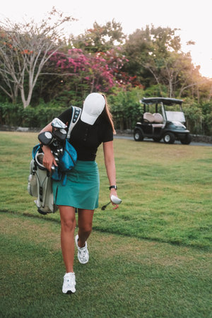 A young woman in a sports skirt walks with a bag containing golf clubs.の写真素材