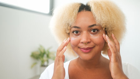 Close up portrait of young African American woman with curly blonde hair with moisturizing patches under her eyes. Skin care in adulthood. Preventing wrinkles and bags under the eyes. Morning routine.の写真素材