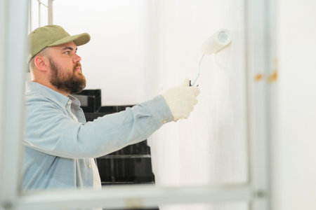 View through a stepladder on a man in construction gloves painting a wall with white paint using a roller. New home renovation, preparing for a housewarming. Professional painter. Worker.の写真素材