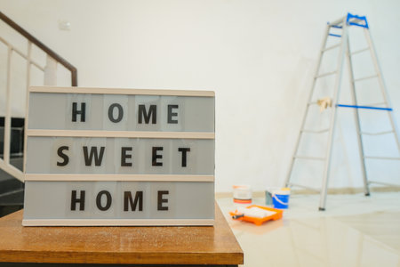 In the foreground is a sign that says Home Sweet Home. In the background, an apartment is being renovated. There is a stepladder next to wall, brushes, cans of paint and putty are lying on the floor.の写真素材