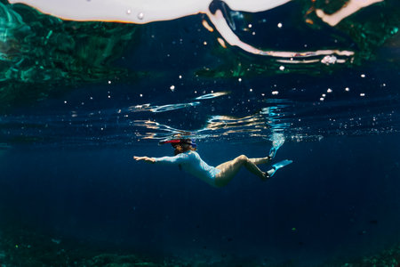 Young woman in blue wetsuit, fins and mask with snorkel swims underwater and looks at coral reef and fish near tropical island. Snorkeling and freediving activity during vacation.の写真素材