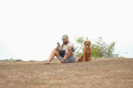 A young man in a cap sits on the grass and looks at his phone. A golden retriever sits next to him, sticking out its tongue and looking into the distance. Walking with a dog in summer.の写真素材