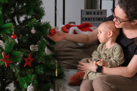 Mom holds her toddler daughter in her pajamas and they look at the Christmas tree together, which is...