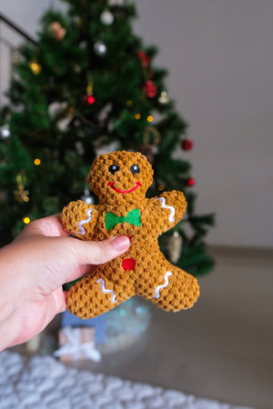 Woman hand holding gingerbread man toy against Christmas tree with decorations and lights. Gift for...