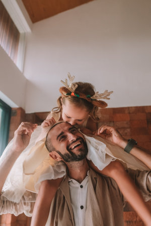 A little girl in a headband with deer antlers and a fluffy dress sits on her dads shoulders. They look at each other and smile happily. Celebrating Christmas with family. Fathers Day.の写真素材