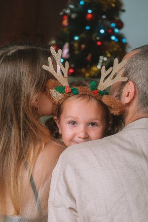 Against the backdrop of Christmas tree, mother, father and their little daughter are hugging, wearing headband with deer antlers. The girl laughs and happily cuddles up to her parents. Family holiday.の写真素材
