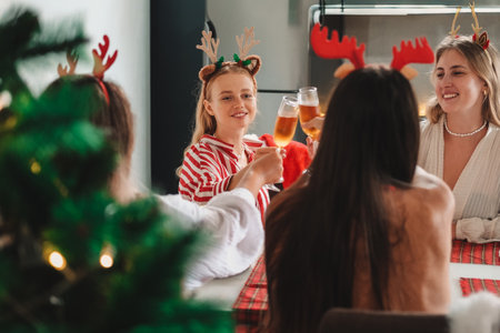 Young women friends wearing headbands with deer antlers are sitting at home at the kitchen table and celebrating Christmas. They clink glasses of champagne and smile. There is a Christmas tree nearby.の写真素材