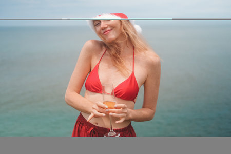 Banner with young red haired woman in Santa hat, red bikini holding glass of champagne standing on...