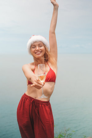 Portrait with young red haired woman in Santa hat, red bikini holding glass of champagne standing on...