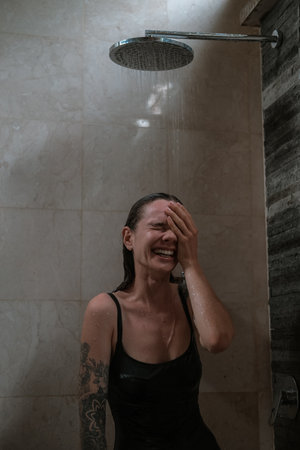 A young woman with wet hair laughs joyfully under a rain shower, covering her face with one hand. She stands in a modern bathroom. The concept happiness, relaxation, and daily self-care.の写真素材