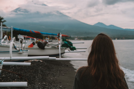 A rear view of a young woman standing on a volcanic beach, facing traditional fishing boats moored...