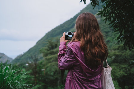 Young woman in purple jacket with brown hair taking pictures of green mountain landscape. She holds...