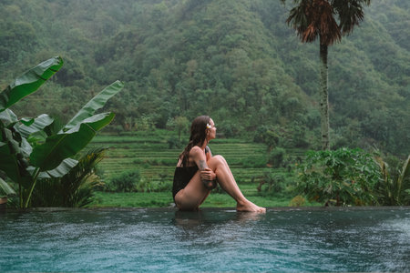 Wide shot a young woman in a black swimsuit sits by an infinity pool, surrounded by tropical plants, overlooking lush green mountains and rice terraces. Eco tourism, travel, nature retreat.の写真素材