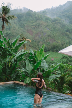 Medium shot of a young woman in a black swimsuit stands in an infinity pool, adjusting her wet hair, surrounded by lush tropical greenery and overlooking misty mountains. Relaxation and self care.の写真素材