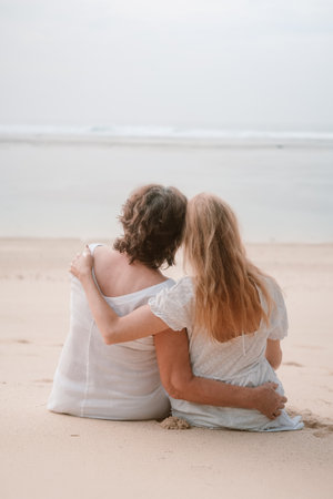 Back view of a senior mother and her young redhead daughter sitting on the beach during a family vacation. They hug and look at the ocean. Mothers Day.の写真素材