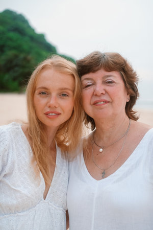 Close up portrait of a young red haired woman and her senior mother standing on the beach by the...