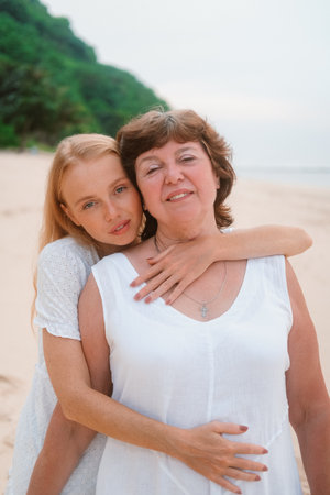 A young redhead charming woman tenderly hugs her senior mother standing on the beach near the ocean during their joint vacation. Daughter and mother on a travel. Mothers day.の写真素材