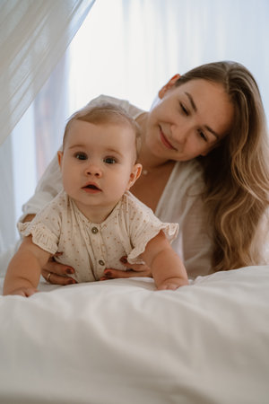 Mother and her baby daughter on a bed on a white blanket. The infant baby is crawling on the bed,...