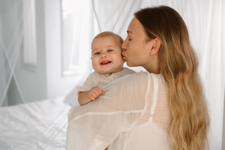 A young woman holds her little daughter and kisses her tenderly. Adorable baby smiles happily. Rocks the baby to sleep. Love and affection.の写真素材