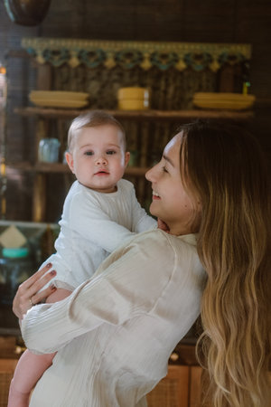 Young mother holding her infant baby daughter talking to her and kissing her forehead in the...