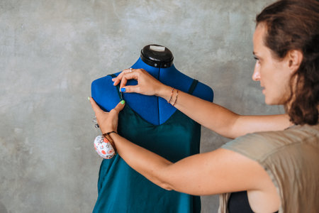 Portrait of young woman fashion designer standing next to mannequin adjusting dress, pinning it with a needle and making chalk marks to re-cut. Small studio atelier. Startup custom clothing store.の写真素材