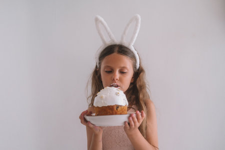 Portrait of a little girl in a rabbit ear headband holding a plate with Easter cake and posing against a white wall in the kitchen. She grimaces, her face is smeared with cream. Spring festive dessertの写真素材