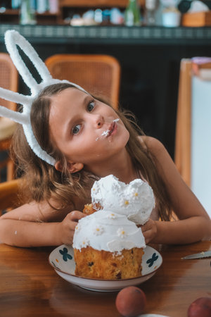 A girl with a rabbit mask on her head eats Easter cake at the kitchen table. She bites a piece of...