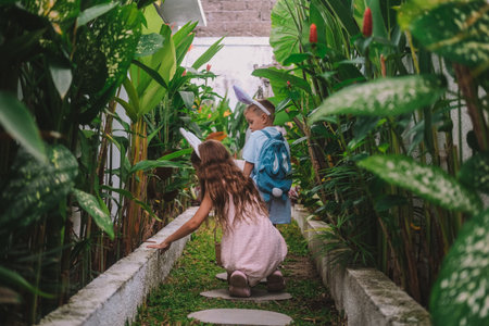 Festive Easter childrens game Egg hunt. Little brother and his older sister run along the path surrounded by tropical greenery in search of eggs, collect them in basket and have fun. Family traditionsの写真素材