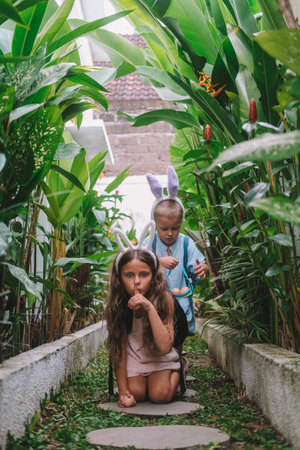 Festive Easter childrens game Egg hunt. Little brother and his older sister run along the path surrounded by tropical greenery in search of eggs, collect them in basket and have fun. Family traditionsの写真素材