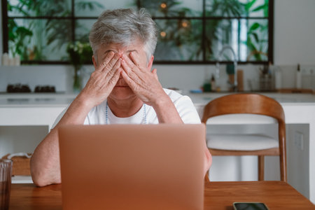 A senior woman with white hair and glasses to improve her vision works on a laptop remotely from...