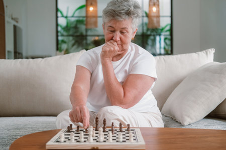 A senior woman with gray hair sits on a sofa in the living room and plays chess on a wooden board. She makes a move and looks ahead at opponent. Leisure and hobbies in retirement. Games for thinking.の写真素材