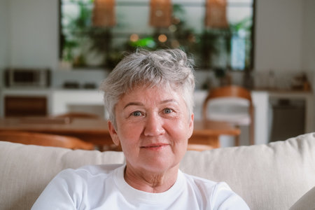 Close portrait of a senior woman with white hair who is sitting on a sofa, she is looking at the camera and starts to smile. Warm smile of a grandmother. Beautiful woman is resting in retirement.の写真素材