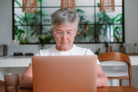 Portrait of a senior woman with gray hair wearing glasses working on a laptop at home in the living room. She is typing on a computer and working remotely from home. Remote work skills for retirees.の写真素材