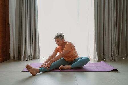 Portrait of an elderly woman with gray hair sitting on a yoga mat in her room and doing exercises. Gymnastics for the elderly and joint support. Improving health in retirement.の写真素材