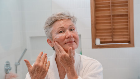 A senior woman stands in the bathroom in front of the mirror with moisturizing patches and applies anti wrinkle cream to face with massage movements. Care for aging skin. Beauty routine in retirement.の写真素材