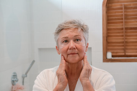 A senior woman with gray hair stands at the mirror with moisturizing patches under her eyes, looks at her reflection and smiles. Care for aging skin and fight against wrinkles. Beauty in retirement.の写真素材