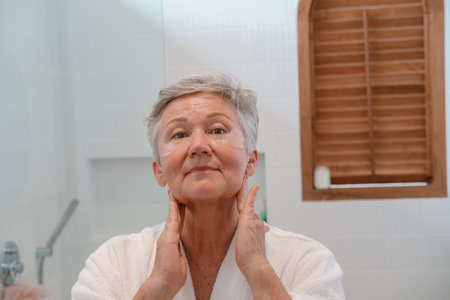A senior woman stands in the bathroom in front of the mirror with moisturizing patches and applies anti wrinkle cream to face with massage movements. Care for aging skin. Beauty routine in retirement.の写真素材