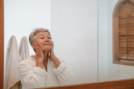 A senior woman stands in the bathroom in front of the mirror and applies moisturizing anti wrinkle cream to her face with massage movements. Care for aging skin. Beauty routine in retirement age.の写真素材