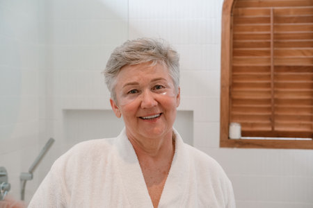 A senior woman with gray hair stands at the mirror with moisturizing patches under her eyes, looks at her reflection and smiles. Care for aging skin and fight against wrinkles. Beauty in retirement.の写真素材
