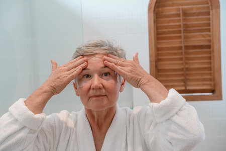 A senior woman stands in the bathroom in front of the mirror with moisturizing patches and applies anti wrinkle cream to face with massage movements. Care for aging skin. Beauty routine in retirement.の写真素材