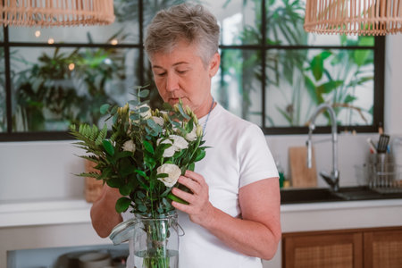 A senior woman with gray hair stands in kitchen next to table on which there is a vase with white roses, she sniffs and adjusts the bouquet.の写真素材