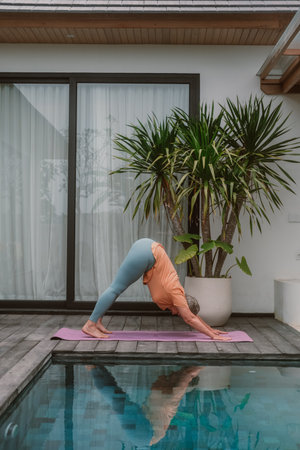 A senior woman with gray hair in a tracksuit does yoga asanas on a mat in a tropical villa with a swimming pool. Health gymnastics for the elderly. Rest and recovery in retirement.の写真素材