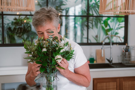 A senior woman with gray hair stands in kitchen next to table on which there is a vase with white roses, she sniffs and adjusts the bouquet.の写真素材