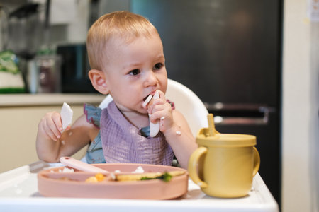 A little girl toddler is sitting on a high chair and eating a cracker. She plays with the cracker imagining that it is an airplane that flies into her mouth and bites is off. The baby plays with foodの写真素材