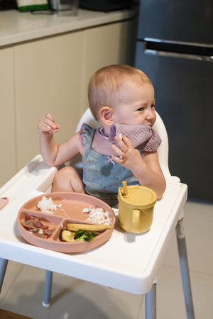 A little toddler girl sits in a high chair and eats her lunch. The baby is learning to eat with a fork, but she picks up rice with her hand and puts it in her mouth. She plays with food and has fun.の写真素材