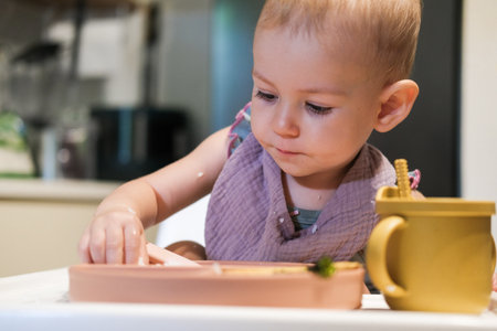 Portrait of little toddler girl sitting in a high chair in the kitchen eating lunch. She thoughtfully puts rice in her mouth with her hand and learns to eat with utensils. The baby eats independently.の写真素材