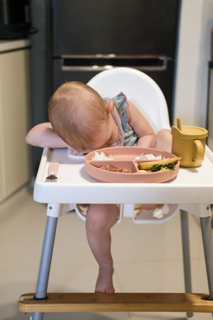 The baby girl does not want to eat and is capricious, sitting on a high chair in the kitchen. The toddler refuses to eat. The child has no appetite. The food is tasteless.の写真素材