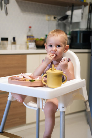 A little girl toddler is sitting on a high chair and eating a cracker. She plays with the cracker imagining that it is an airplane that flies into her mouth and bites is off.の写真素材