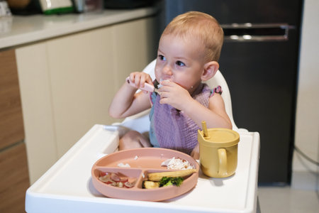 Portrait of little toddler girl sitting in a high chair in the kitchen eating lunch. She thoughtfully puts rice in her mouth with her hand and learns to eat with utensils. The baby eats independently.の写真素材
